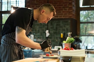 Caucasian young adult man wearing chef uniform preparing gourmet dish in professional kitchen, carefully plating food with gloved hands, fresh vegetables visible on counter