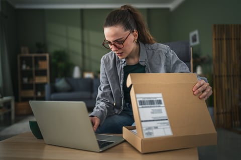 Woman in glasses opening a cardboard box while looking at a laptop
