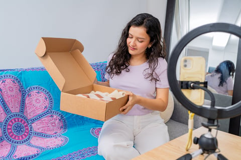 Young female influencer recording video for social media followers, unboxing clothes received in a cardboard package