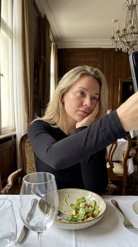 Woman in black long-sleeved shirt taking selfie at dining table with salad and wine glass in elegant room with chandelier