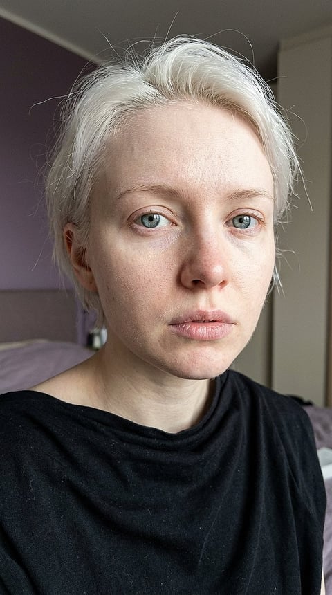 Close-up portrait of a person with short platinum blonde hair and light blue eyes, wearing a black shirt, taken indoors