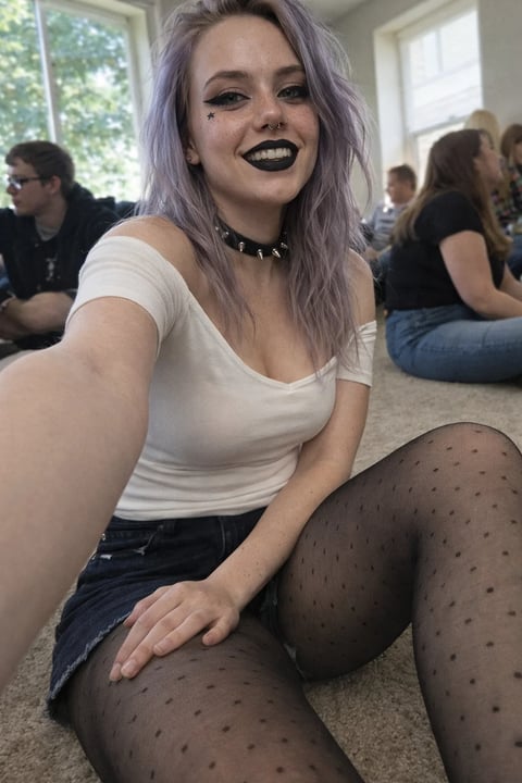 Woman with lavender hair, dark lipstick, white shirt, and polka dot tights smiling at camera in casual indoor setting with others in background