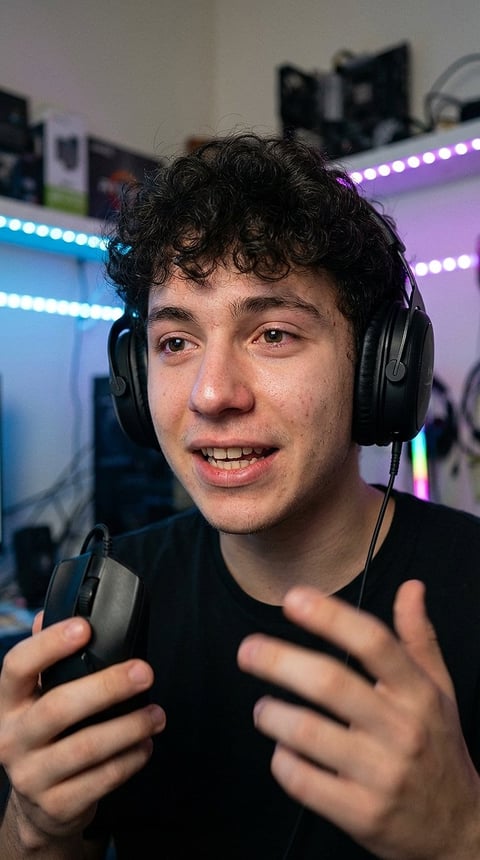 Young man wearing black headphones in a gaming setup with LED lights, smiling and gesturing with his hands