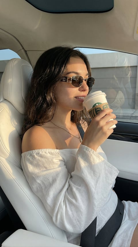 Woman wearing sunglasses and off-shoulder white shirt sitting in a car seat, holding a coffee cup