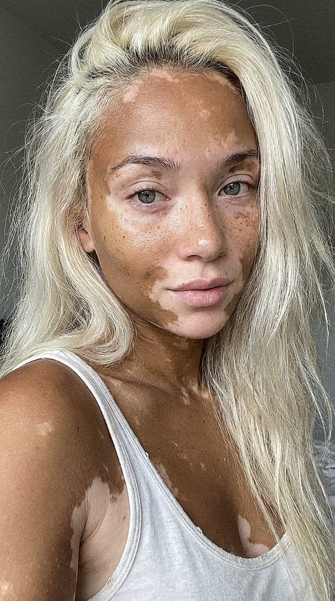 Portrait of a woman with platinum blonde wavy hair, freckles, and light eyes wearing a white tank top against a neutral gray background